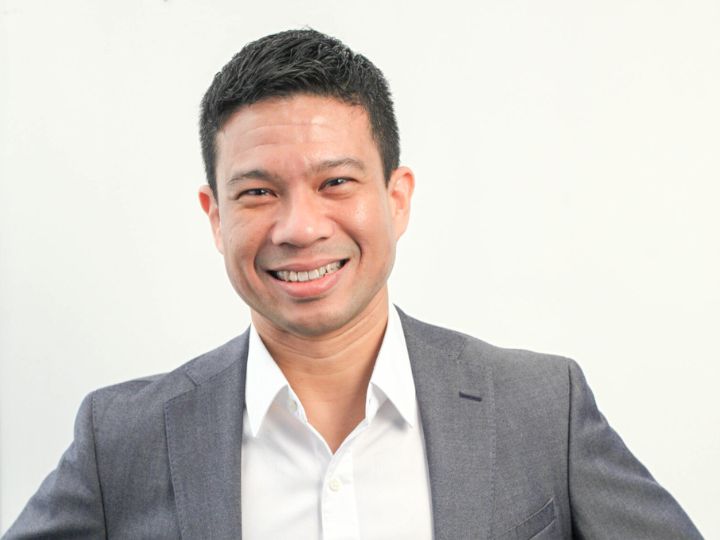 Un joven con cabello negro corto, camisa blanca, saco azul marino y gafas sonríe a la cámara.