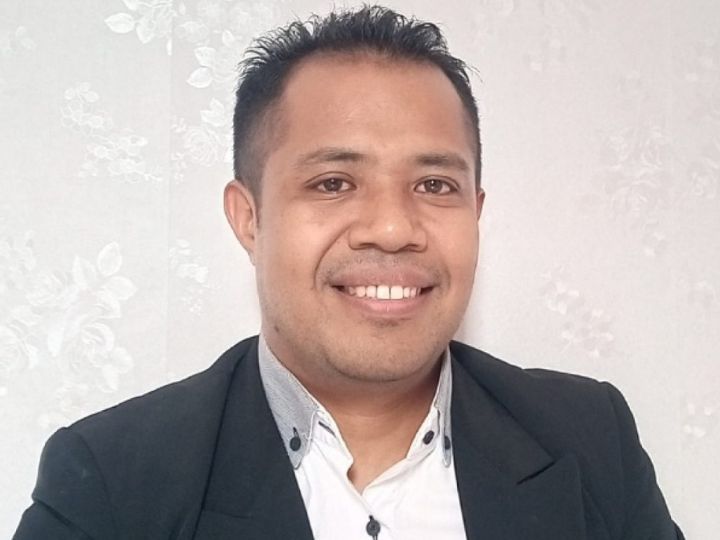 Un hombre joven con la cabeza rapada y barba corta, con gafas y una chaqueta gris oscuro sobre una camiseta roja, levanta la mano derecha mientras habla.